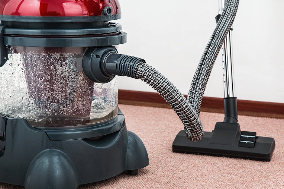A person wearing beige sneakers and blue jeans is operating a vacuum cleaner with a yellow body and black hose, cleaning a traditional patterned carpet in a residential setting. The carpet features an
