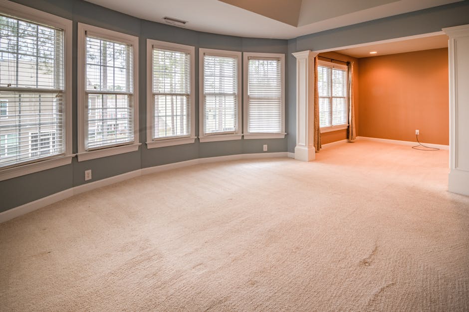 A spacious, well-lit living room featuring light beige carpet flooring that appears clean and free of stains. The room has multiple large double-hung windows with white wooden blinds, allowing natural
