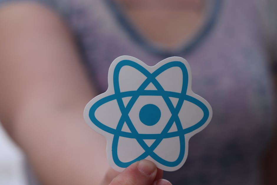 A person holding a sticker with a blue atom symbol in front of their chest, which appears to be outdoors or in a well-lit indoor environment. The individual is wearing a grey sleeveless top, and the b